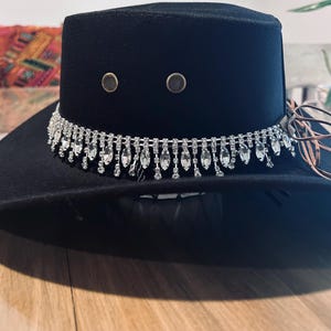 May include: A black fedora hat with a decorative silver rhinestone band. The hat features two small, round, metallic accents on the crown. The brim is wide, and the hat appears to be made of a soft material. A stylish accessory.