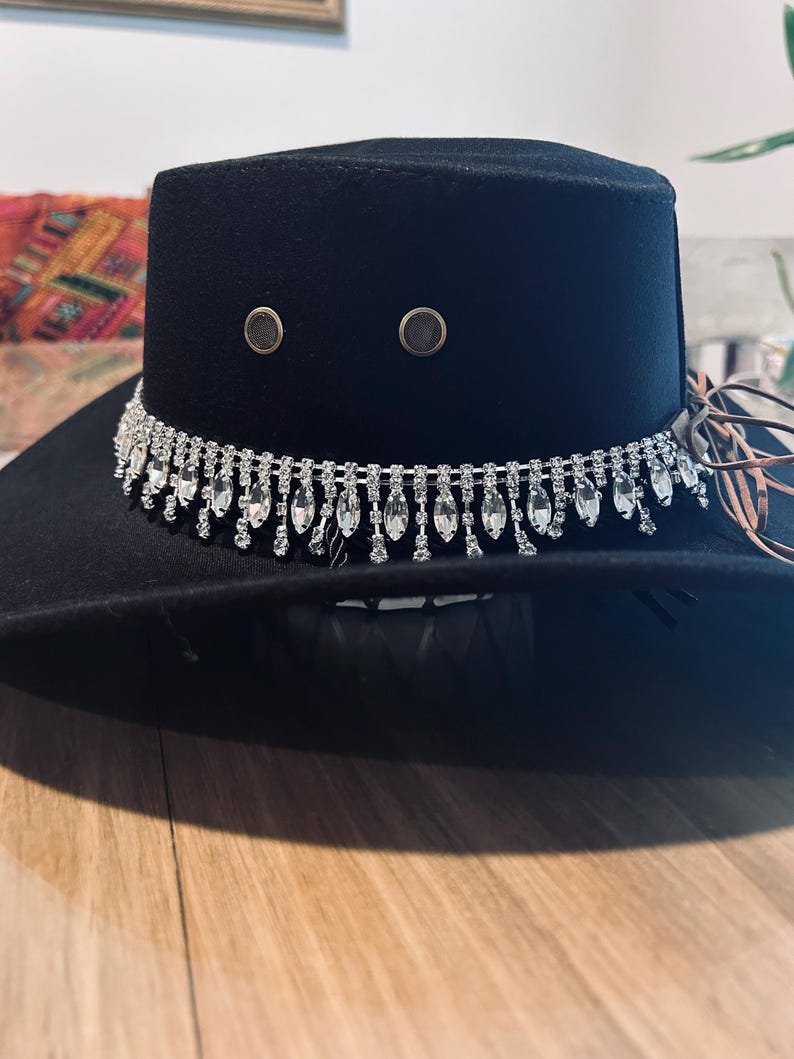 May include: A black fedora hat with a decorative silver rhinestone band. The hat features two small, round, metallic accents on the crown. The brim is wide, and the hat appears to be made of a soft material. A stylish accessory.