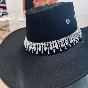 May include: Black cowboy hat with a decorative silver rhinestone band around the crown. The hat has a wide brim and a structured crown with three silver eyelets. The hat is made of a felt-like material.