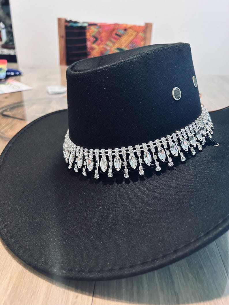 May include: Black cowboy hat with a decorative silver rhinestone band around the crown. The hat has a wide brim and a structured crown with three silver eyelets. The hat is made of a felt-like material.
