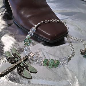 May include: A dragonfly brooch and necklace with a brown boot on a silver surface. The necklace features clear and green beads. The brooch is light green with black accents and clear rhinestones. The necklace has a dragonfly charm.