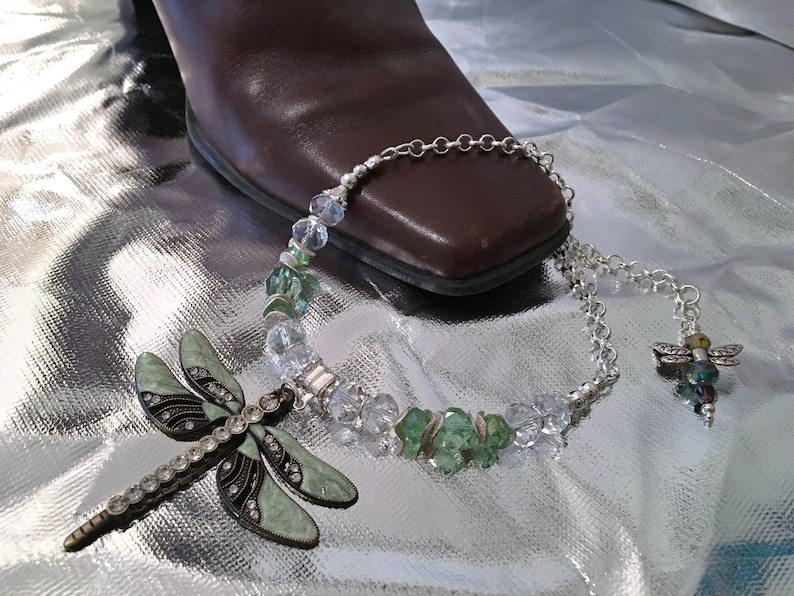 May include: A dragonfly brooch and necklace with a brown boot on a silver surface. The necklace features clear and green beads. The brooch is light green with black accents and clear rhinestones. The necklace has a dragonfly charm.