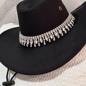 May include: A black cowboy hat with a sparkling rhinestone band around the crown. The hat has a wide brim and a black chin strap. The hat is made of a soft material.