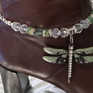 May include: A brown leather boot adorned with a decorative anklet. The anklet features clear and green beads, with a dangling dragonfly charm. The dragonfly has green wings, black accents, and a silver-colored body.