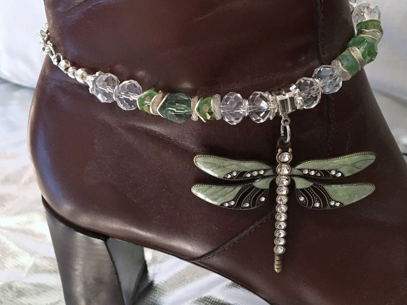 May include: A brown leather boot adorned with a decorative anklet. The anklet features clear and green beads, with a dangling dragonfly charm. The dragonfly has green wings, black accents, and a silver-colored body.