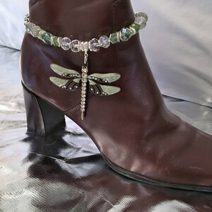 May include: A brown leather ankle boot with a decorative dragonfly anklet. The anklet features clear and green beads with a dangling dragonfly charm. The dragonfly has pale green wings and a rhinestone-studded body.
