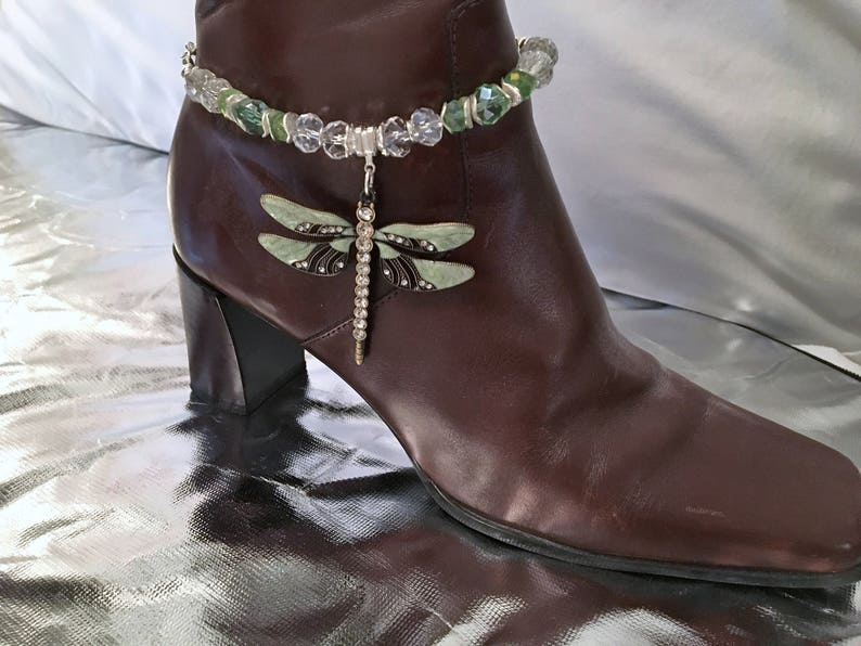 May include: A brown leather ankle boot with a decorative dragonfly anklet. The anklet features clear and green beads with a dangling dragonfly charm. The dragonfly has pale green wings and a rhinestone-studded body.