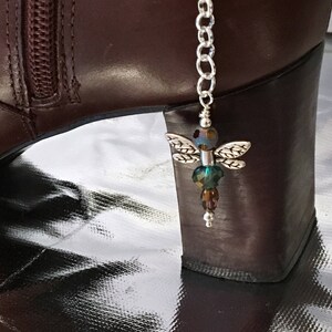 May include: A silver dragonfly boot charm with iridescent beads and detailed wings hangs from a silver chain. The charm is attached to a brown leather boot with a zipper.
