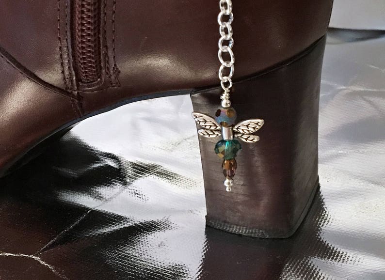 May include: A silver dragonfly boot charm with iridescent beads and detailed wings hangs from a silver chain. The charm is attached to a brown leather boot with a zipper.
