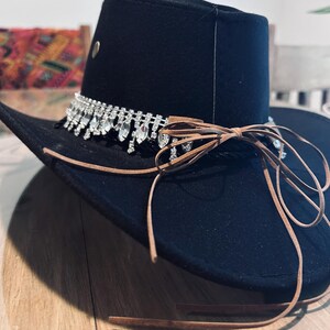 May include: Black cowboy hat with a decorative rhinestone band and brown leather ties. The hat has a wide brim and a structured crown. A small metal stud is visible on the side of the crown. The hat is likely made of felt or a similar material.