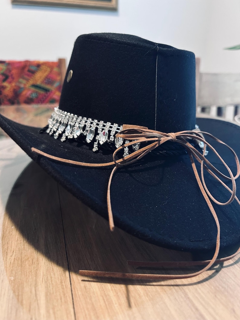 May include: Black cowboy hat with a decorative rhinestone band and brown leather ties. The hat has a wide brim and a structured crown. A small metal stud is visible on the side of the crown. The hat is likely made of felt or a similar material.