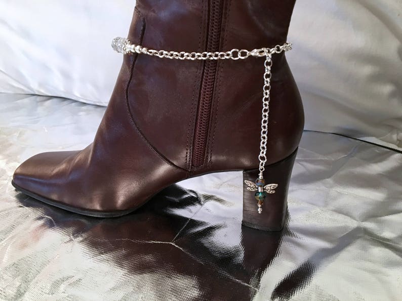 May include: A silver-toned boot chain with a dragonfly charm is draped over a brown leather boot. The chain features a series of small, clear beads and a dangling dragonfly pendant with iridescent accents. The boot has a square toe and a small heel.