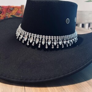 May include: Black cowboy hat with a decorative rhinestone band around the crown. The hat has a wide brim and a structured crown. Two silver-colored eyelets are visible on the side of the crown. The hat is sitting on a wooden surface.