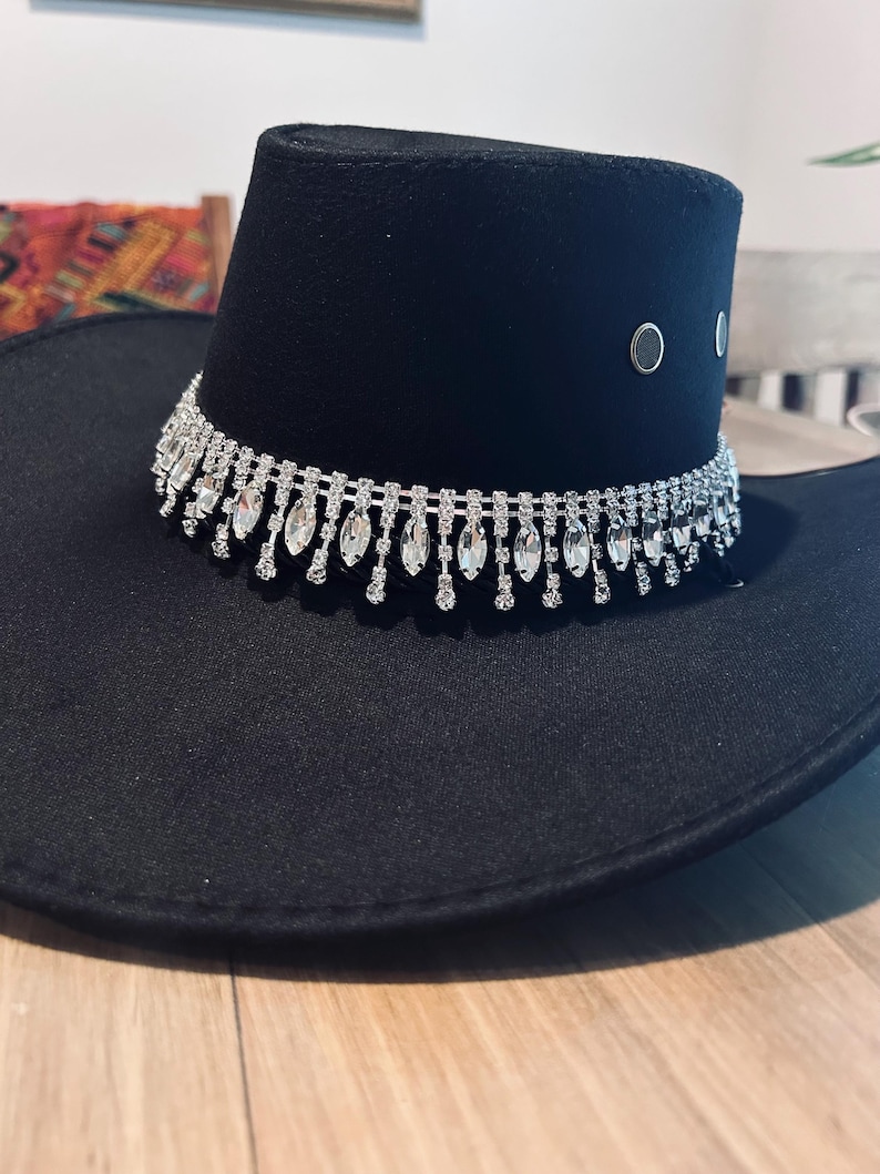 May include: Black cowboy hat with a decorative rhinestone band around the crown. The hat has a wide brim and a structured crown. Two silver-colored eyelets are visible on the side of the crown. The hat is sitting on a wooden surface.