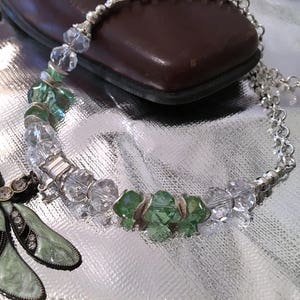 May include: A silver necklace with clear and green crystal beads. A decorative dragonfly pendant with green enamel and rhinestones is on the left. The necklace is on a reflective surface.