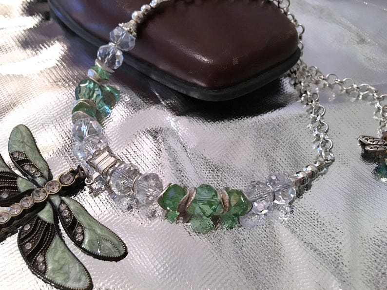 May include: A silver necklace with clear and green crystal beads. A decorative dragonfly pendant with green enamel and rhinestones is on the left. The necklace is on a reflective surface.