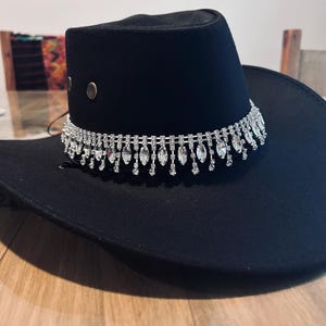 May include: A black cowboy hat with a decorative silver rhinestone band. The hat has a wide brim and a rounded crown. Two silver rivets are on the side of the crown.