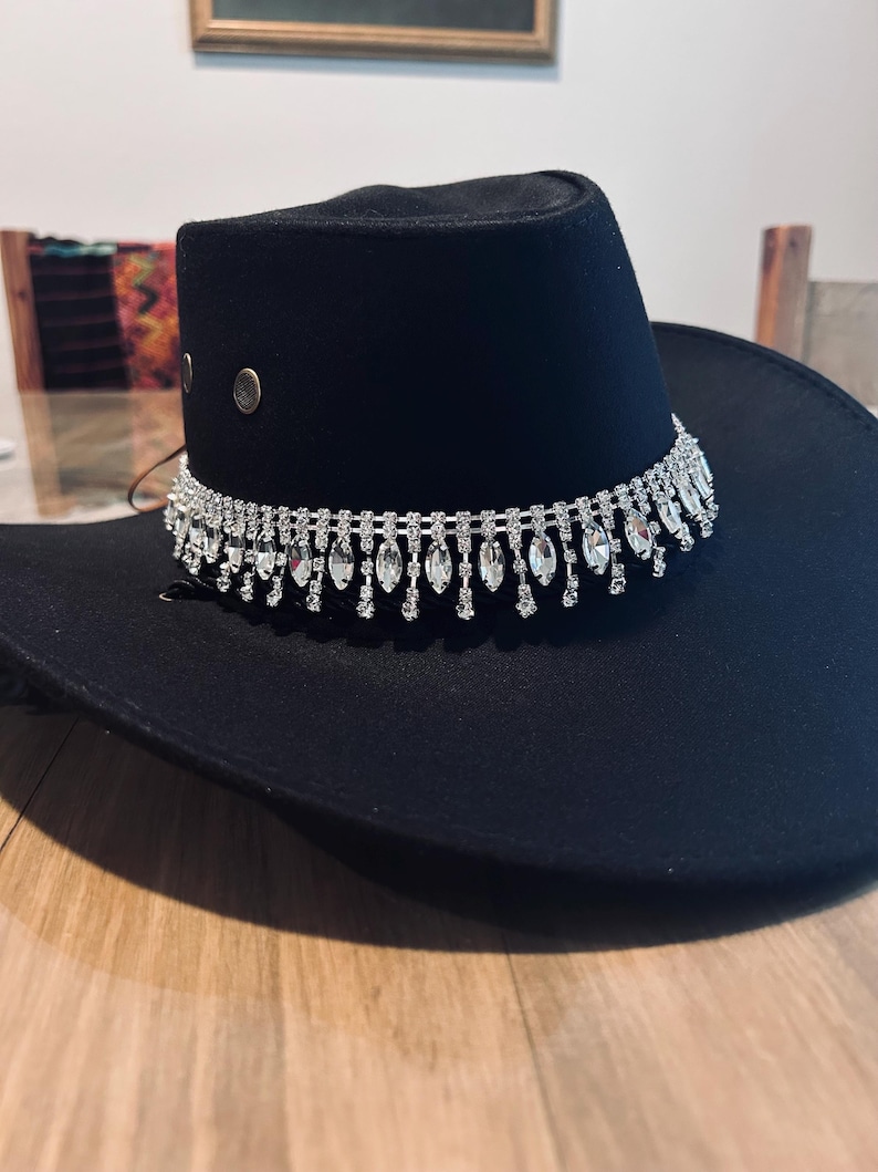 May include: A black cowboy hat with a decorative silver rhinestone band. The hat has a wide brim and a rounded crown. Two silver rivets are on the side of the crown.