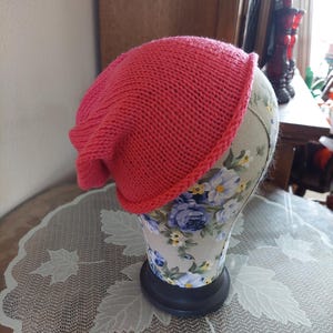 May include: A vibrant pink knitted beanie hat. The hat is displayed on a floral patterned mannequin head. The hat has a folded brim and a slightly slouchy fit. The hat is made of a soft, textured yarn.