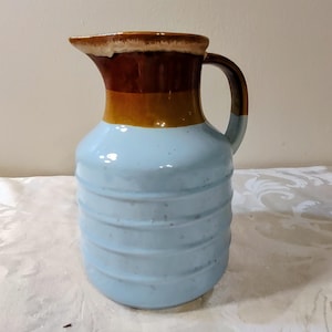 May include: A ceramic pitcher with a light blue body and a brown, tan, and white rim. The pitcher has a handle and a ridged design around the body. This decorative pitcher is suitable for serving beverages.
