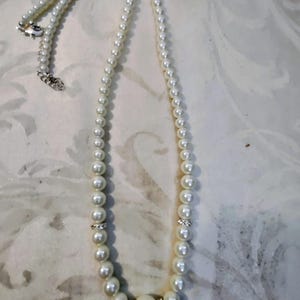 May include: A cream-colored pearl necklace with a silver clasp. The necklace features graduated pearls, with larger pearls at the center and smaller ones towards the clasp. Small, clear crystals are set between some of the pearls.