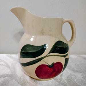 May include: Cream-colored ceramic pitcher with a handle and spout. The pitcher is decorated with a red apple and green leaf design. The pitcher is likely used for serving beverages.