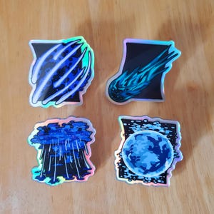 May include: Four holographic stickers with celestial designs. The stickers feature a blue meteor, a blue planet, a blue rain cloud, and a blue and black abstract design. Each sticker has a black background and a silver border.
