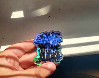 Puede incluir: Una pegatina holográfica con una ilustración estilizada de una nube de lluvia. El diseño incluye un cielo azul oscuro con estrellas blancas, rayas de lluvia oscuras y un borde iridiscente. La pegatina se sostiene en una mano.