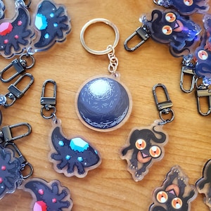 May include: A collection of acrylic keychains with various designs. One keychain features a round, dark blue planet design. Other keychains have black creature designs with colorful gem accents and metal clasps.
