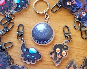May include: A collection of acrylic keychains with various designs. One keychain features a round, dark blue planet design. Other keychains have black creature designs with colorful gem accents and metal clasps.