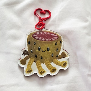 May include: A clear acrylic keychain featuring a cartoon monster with a smiling face, multiple eyes, and tentacle-like legs. The monster is primarily brown with a red mouth and is attached to a red heart-shaped clasp.