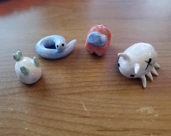May include: Four small, hand-sculpted clay figurines. One is a white creature with green accents, one is a light blue coiled worm, one is a rounded red figure with a blue visor, and one is a white multi-legged creature with black eyes and markings.