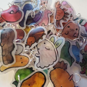 Puede incluir: Una colección de dijes coloridos pintados a mano con varios diseños, incluyendo un fantasma, un escarabajo, un camarón, un dinosaurio y un hongo. Los dijes están hechos de plástico transparente y tienen anillos de salto plateados.