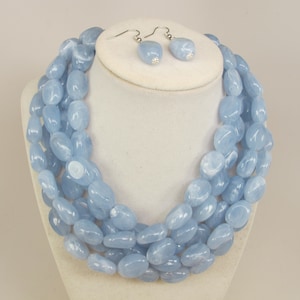 May include: A light blue beaded necklace and matching earrings. The necklace has multiple strands of oval-shaped beads. The earrings have a similar design, with a single bead suspended from a silver hook. The jewellery is displayed on a white bust.