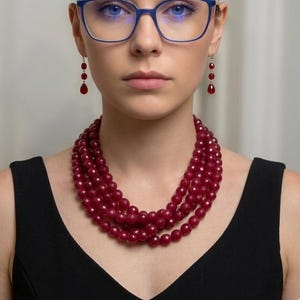 May include: A woman wearing a multi-strand red beaded necklace, matching earrings, and blue-framed glasses. The necklace is layered and sits close to the neck. The earrings have a dangling, teardrop design. The woman is wearing a black top.