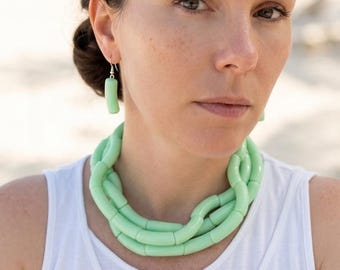 May include: A woman wearing a light green necklace and matching earrings. The necklace is made of rounded, tube-shaped beads. She is wearing a white tank top. The background is a beach with the ocean in the distance.