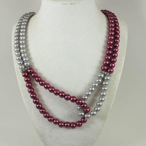 Peut inclure: Un collier à deux rangs avec des sections alternées de fausses perles argentées et bordeaux. Les rangs sont reliés par des barres argentées ornées de petits strass transparents. Le collier est présenté sur un buste de mannequin blanc.