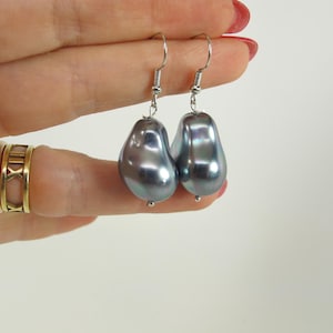 May include: A pair of teardrop-shaped, iridescent gray pearl earrings with silver-colored hooks. The earrings are held up by a hand with gold rings on the fingers. The pearls have a smooth, lustrous surface.