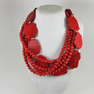 May include: A multi-strand necklace featuring vibrant red spherical beads interspersed with large, flat, irregular red stone slabs. Small silver-toned metal spacers are placed between the beads and the larger stone pieces throughout the design.