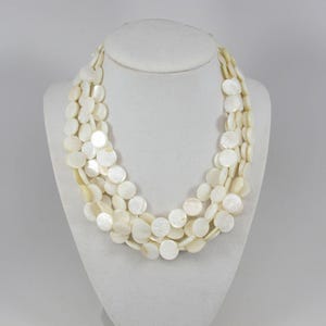 May include: A multi-strand necklace composed of numerous small, circular, off-white mother-of-pearl shell discs. The discs are arranged in several overlapping layers, creating a textured, layered appearance with a subtle iridescent sheen.