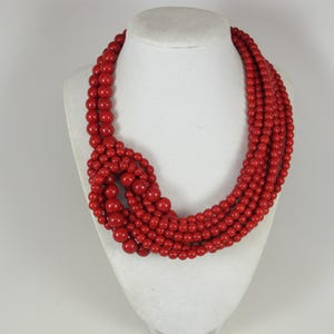 May include: A bold red beaded necklace displayed on a white mannequin bust. The necklace features multiple strands of round, vibrant red beads, knotted together in a unique design. The beads vary in size, adding visual interest to the jewelry.