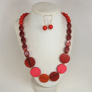 May include: A red beaded necklace and matching earrings set. The necklace features a variety of red beads, including round, oval, and disc-shaped elements. The earrings have a single red bead. The jewelry is displayed on a white mannequin.