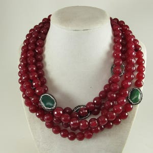 Puede incluir: Un collar de múltiples hilos con cuentas de color rojo intenso facetadas dispuestas en varias capas. El diseño está acentuado con tres piedras verdes lisas de forma ovalada, cada una enmarcada por un borde de pequeños cristales claros engastados en metal oscuro.