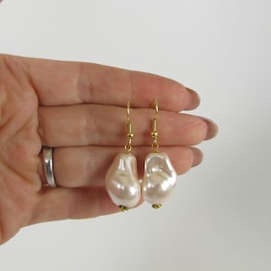 May include: A pair of pearl-colored, baroque-style earrings with gold-colored hooks and small gold-colored beads at the base of each earring. The earrings are held against a white background.