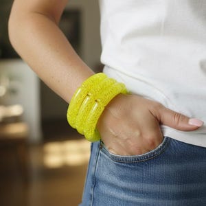 May include: A stack of three bright yellow bracelets. The bracelets are made of a textured material and are worn on the wrist. The person is wearing a white t-shirt and blue jeans.