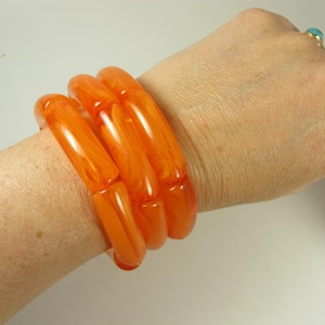 May include: A set of four orange, chunky, translucent bracelets. The bracelets are worn on a wrist, showcasing their vibrant color and smooth, rounded design. The bracelets appear to be made of a resin material.