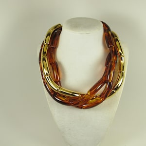 May include: A layered necklace with amber-colored, barrel-shaped beads and gold-tone metal accents. The necklace is displayed on a white bust form, showcasing its design and color contrast. The jewelry is a statement piece.
