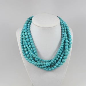 May include: A multi-strand necklace featuring turquoise-colored beads of varying sizes. The necklace is displayed on a white mannequin bust, showcasing its layered design and vibrant color. The beads have a smooth, rounded shape.
