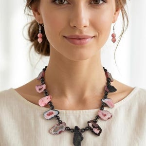 May include: A necklace featuring irregular, polished slices of pink and black agate stones interspersed with small, round, dark metallic beads, paired with matching drop earrings consisting of two small pink spherical beads on silver hooks.