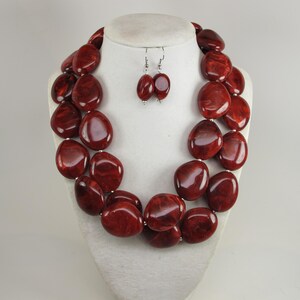 May include: A two-strand necklace and matching earrings set. The jewelry features large, oval-shaped beads in a deep red color with a marbled effect. The necklace and earrings are displayed on a white mannequin bust.